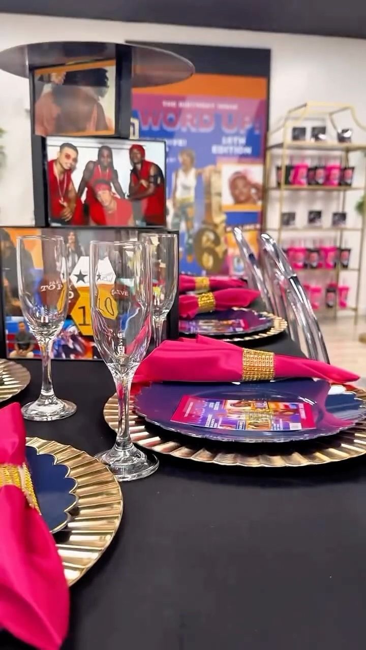 Table setting with gold-rimmed plates, wine glasses, and bright pink napkins on a black tablecloth.