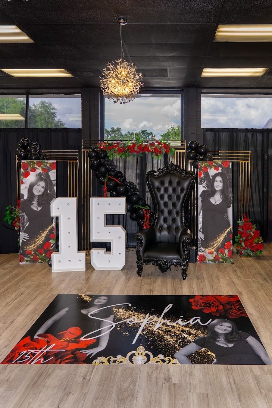 Black-and-red party display with large “15” balloons, throne chair, and decorated backdrop indoors