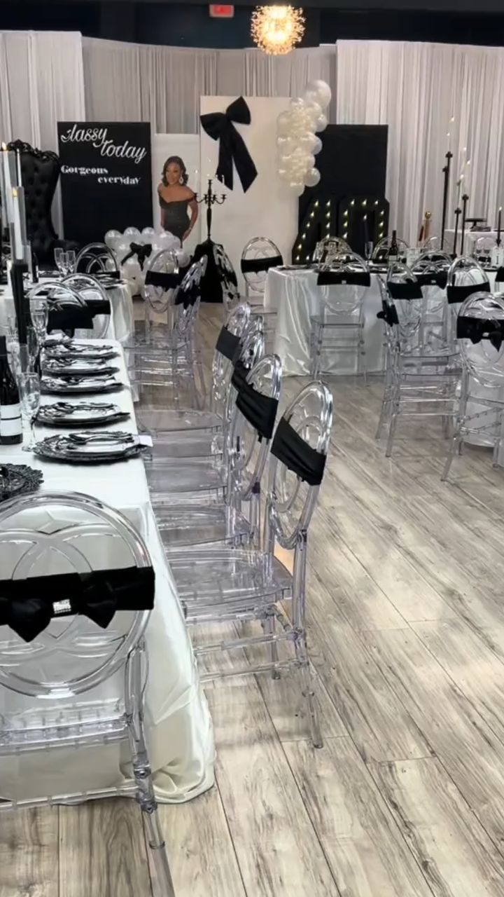 Decorated banquet hall with black-and-white tables, clear chairs, and white draped backdrops