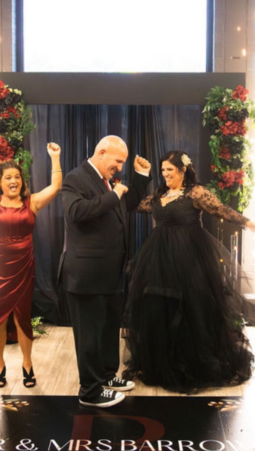 Three people posing on a decorated stage, one speaking into a microphone, with floral arches and curtains behind them.