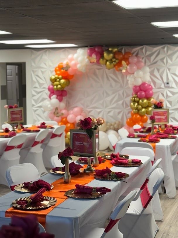 Decorated banquet hall with white chairs, orange table settings, and a pink-and-gold balloon backdrop