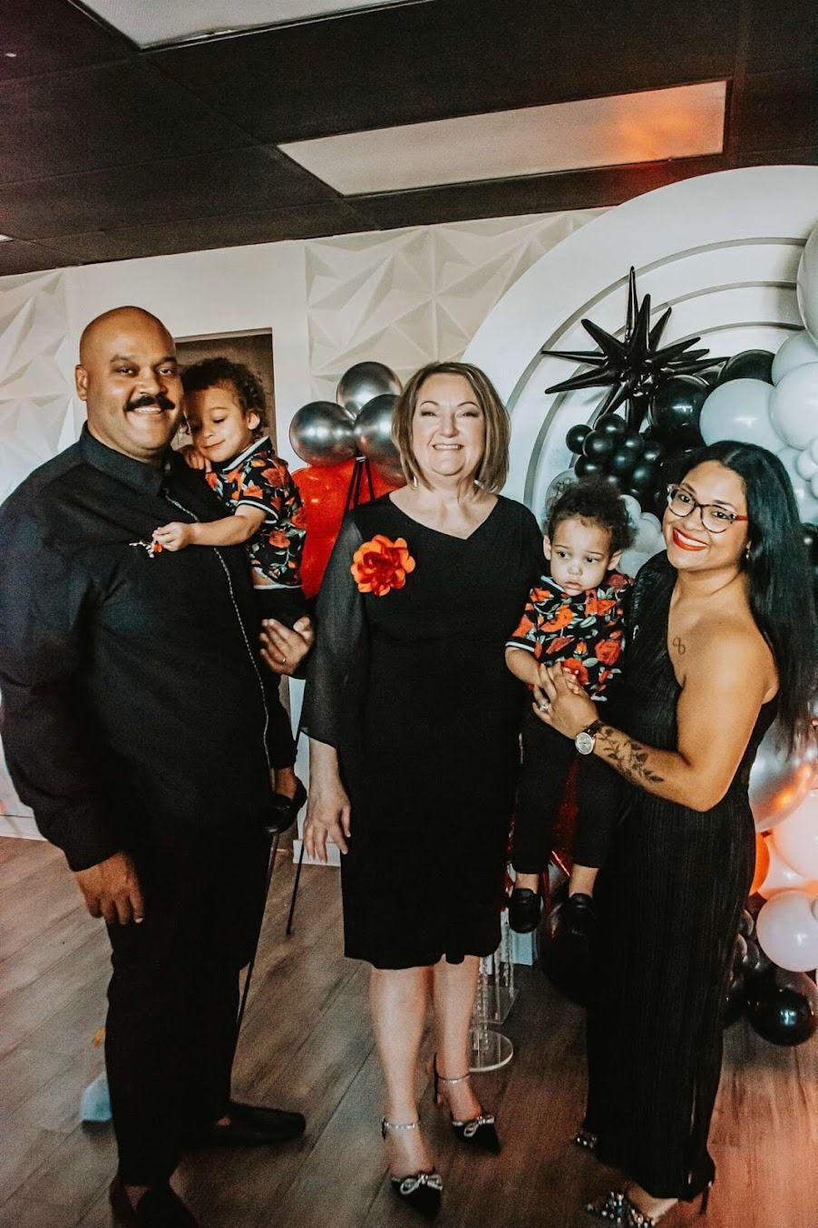Four people posing at a party, with black, red, and silver balloon decorations in the background.