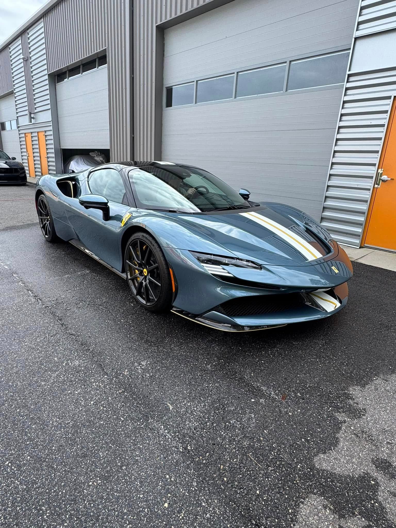 Blue Ferrari SF90 supercar parked in front of a building with a wet asphalt surface.