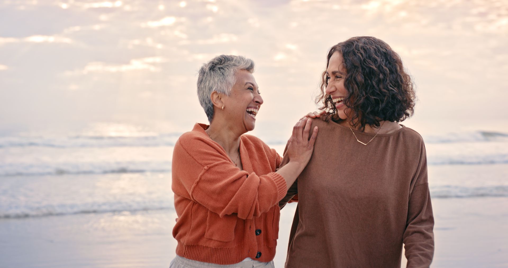 Two women smiling, one has arm around the other's shoulder on a beach at sunset.
