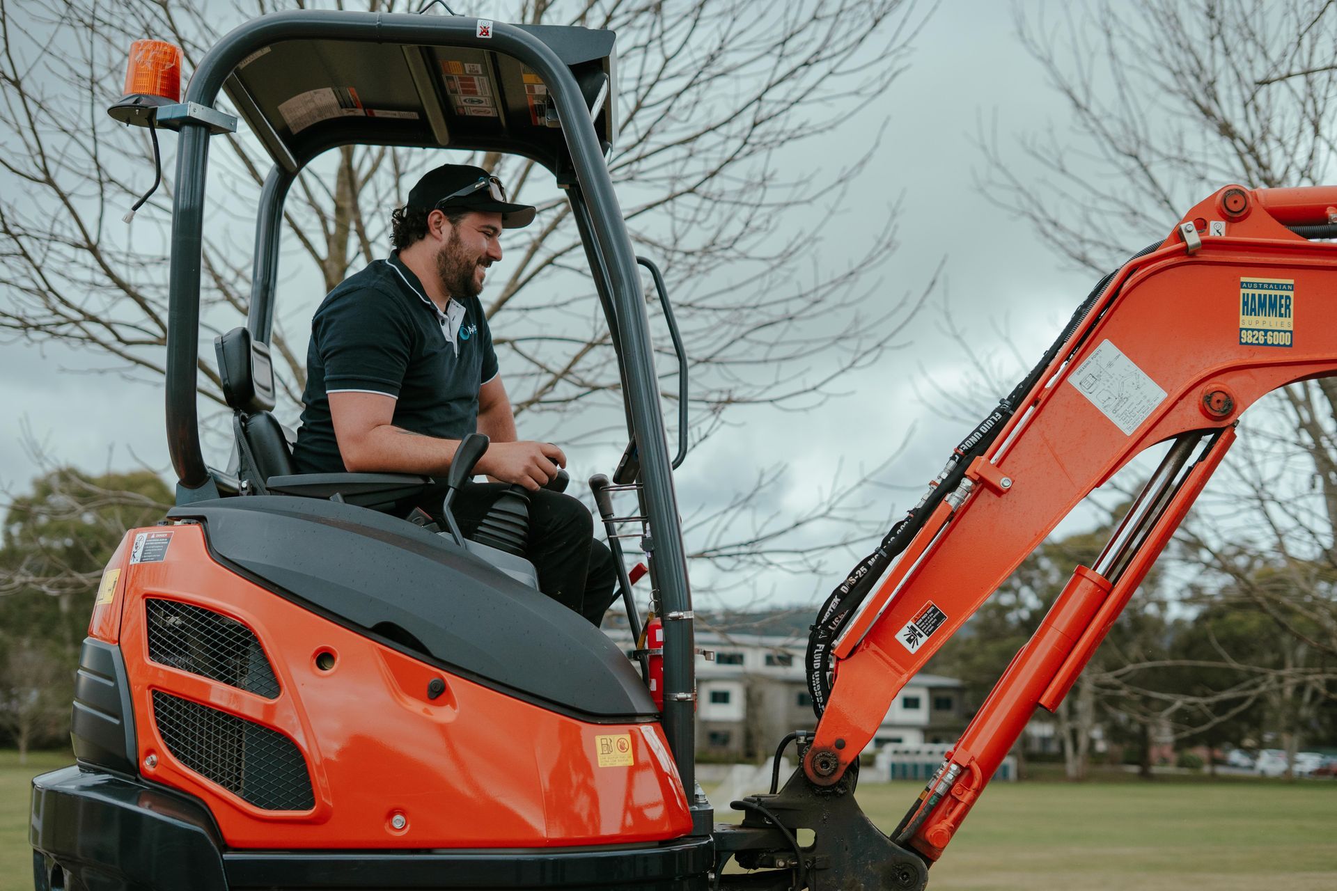 Man sitting in an orange excavator cab outdoors, with the boom extended beside him. — Aquafix Plumbing Services in Bowral, NSW