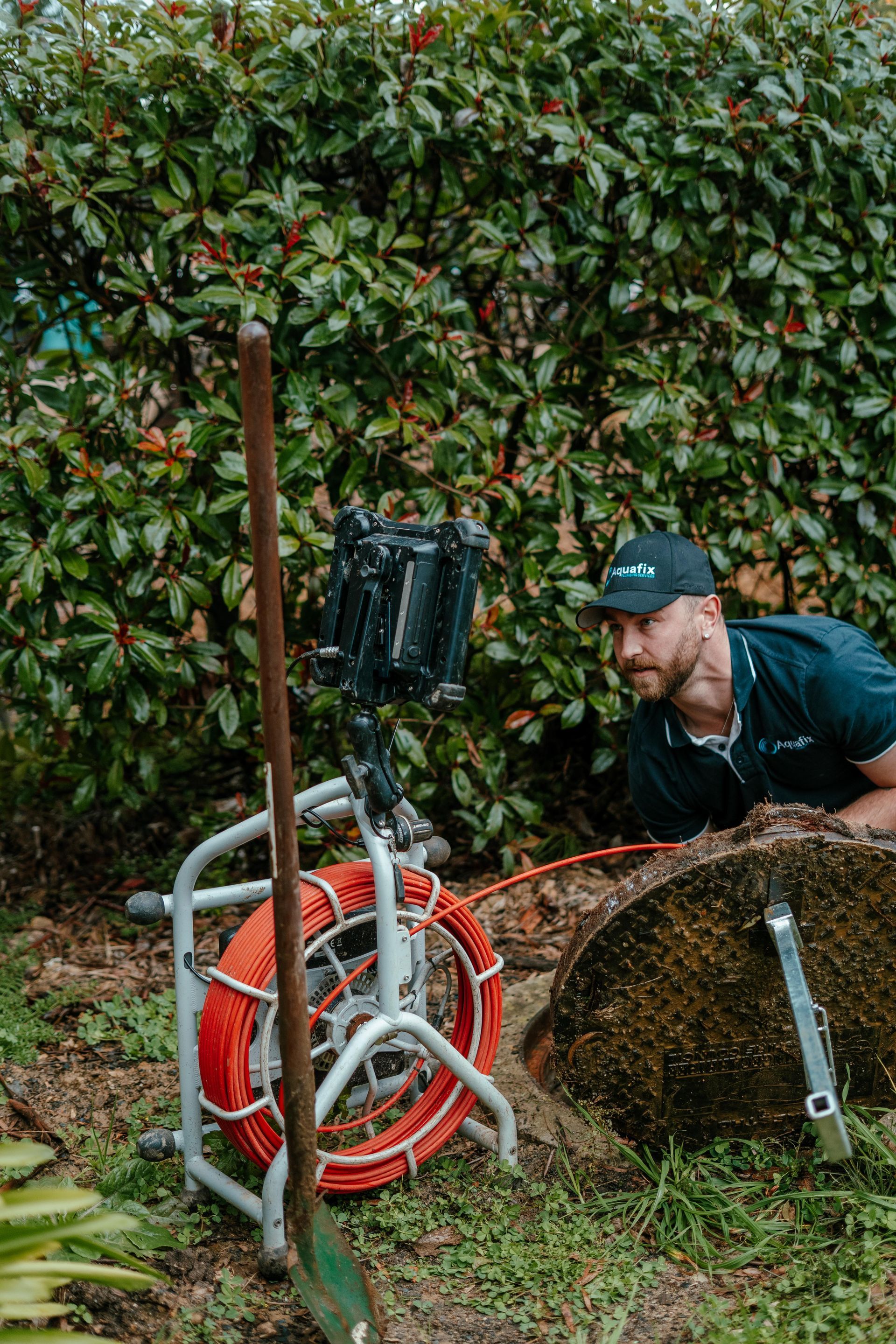 Worker crouches beside a hedge with a hose reel and tools in a garden pit. — Aquafix Plumbing Services in Bowral, NSW
