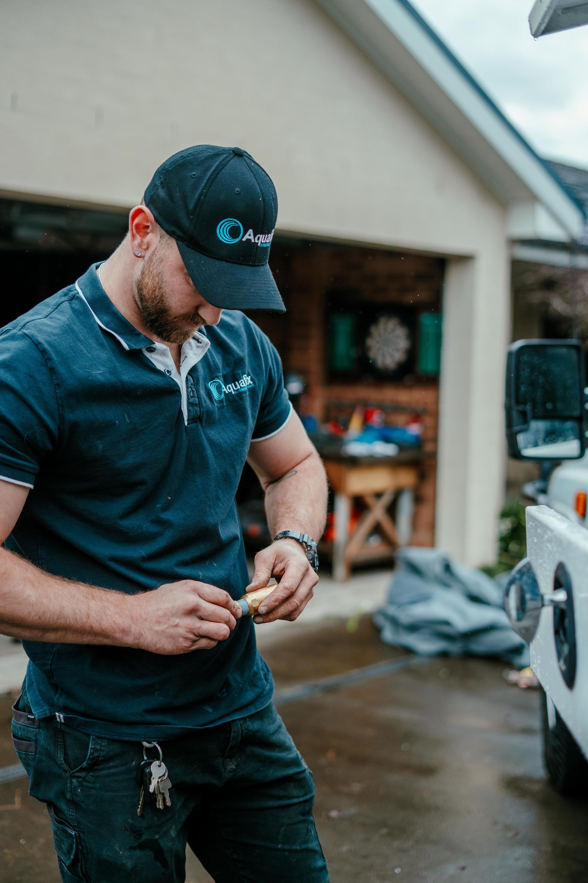 Man in a dark cap and shirt working on a vehicle in a wet driveway near a garage — Aquafix Plumbing Services in Bowral, NSW