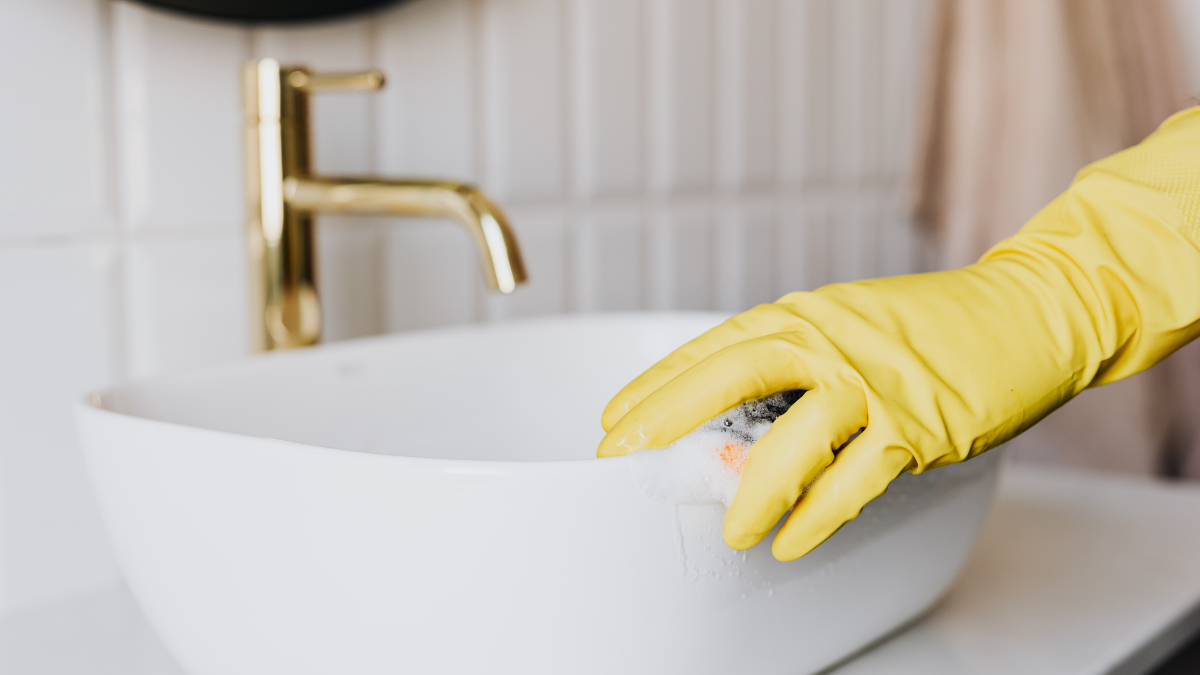 Hand in yellow glove cleans a white sink in a bathroom; gold faucet visible.