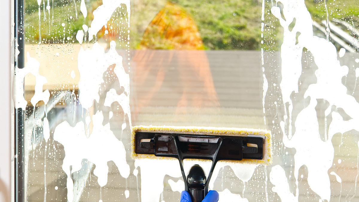 A person cleans a window with a soapy mop, removing foam; outside, a blurred view of trees.
