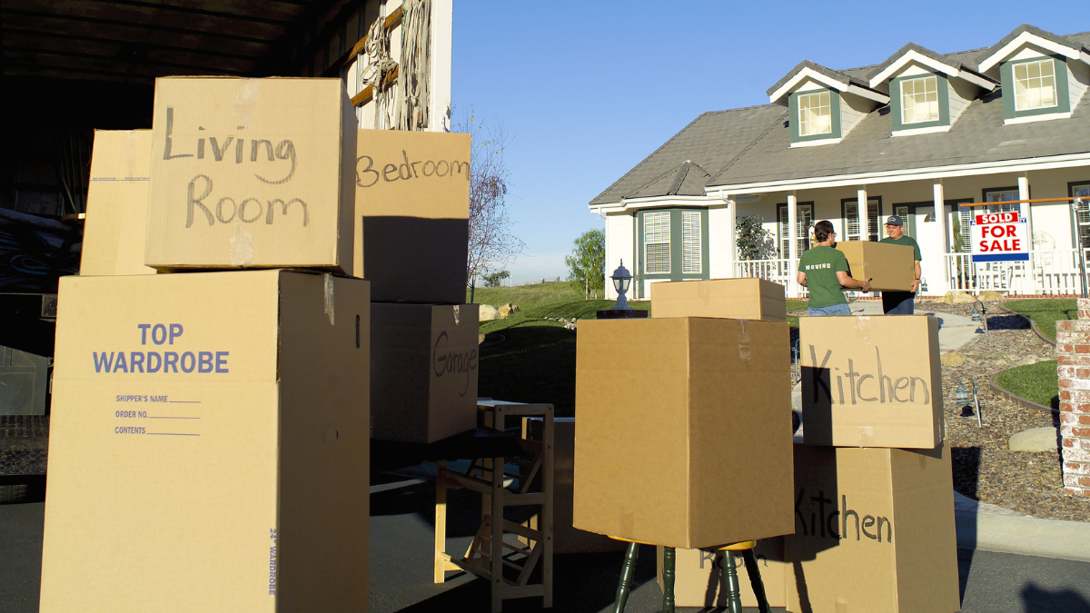 Cardboard boxes for moving are stacked in a driveway, with people carrying boxes toward a house with a 
