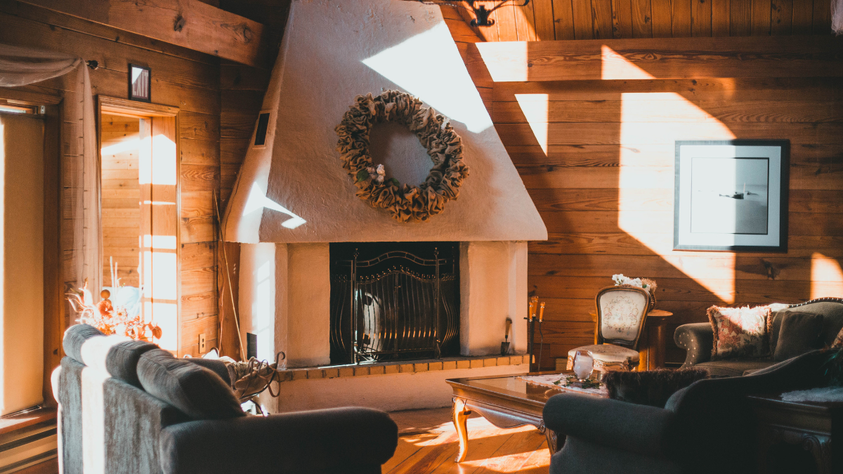Cozy living room with fireplace, wooden walls, sunlight. A wreath hangs above the fireplace.