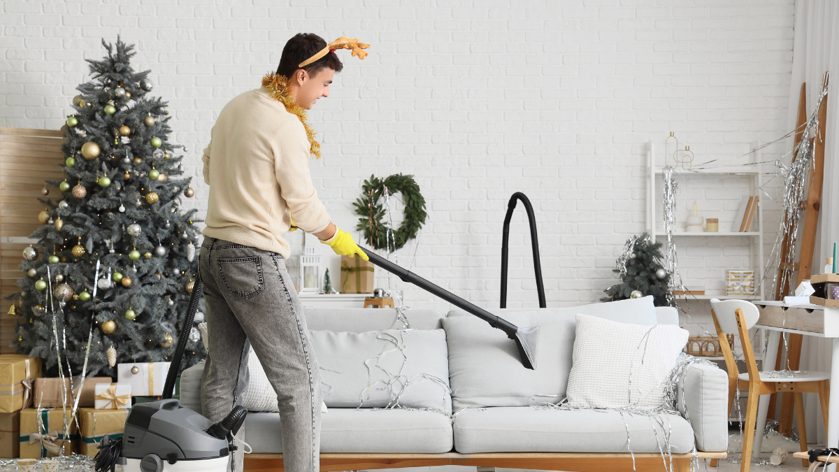 A person wearing reindeer antlers uses a vacuum cleaner to tidy a light gray sofa in a room with a decorated Christmas tree.