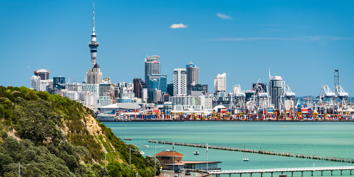 A scenic view of the Auckland skyline, including the Sky Tower, along a vibrant waterfront under a clear blue sky.