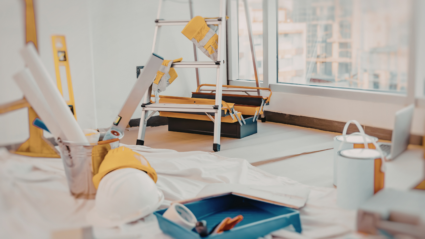 Person painting at a bright desk with art supplies, sketchbook, and a lamp near a window.