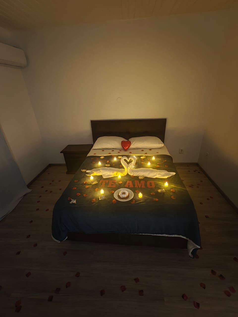 Quarto com cama arrumada, decorada com luzinhas brilhantes e uma almofada em forma de rosa.