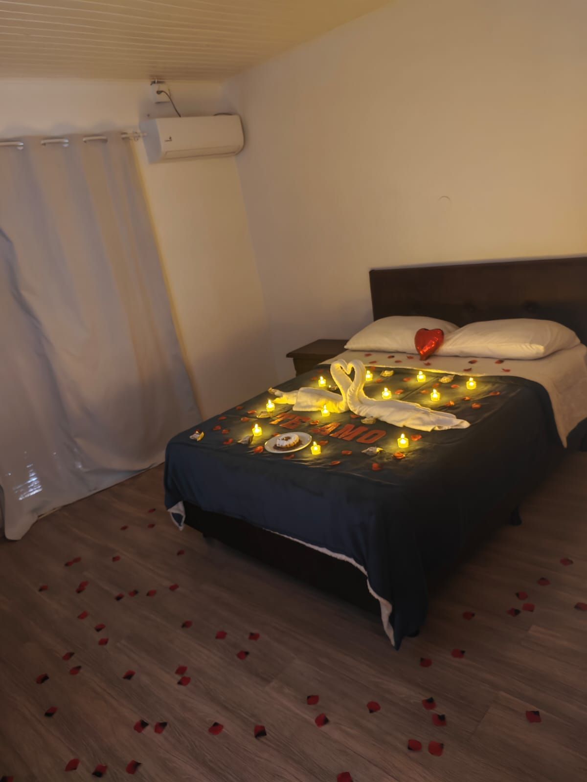 Quarto com cama decorada com luzes, uma almofada em formato de coração vermelho e pétalas de rosa no chão.