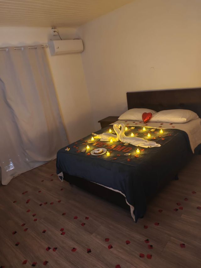 Quarto com cama decorada com luzes, uma almofada em formato de coração vermelho e pétalas de rosa no chão.