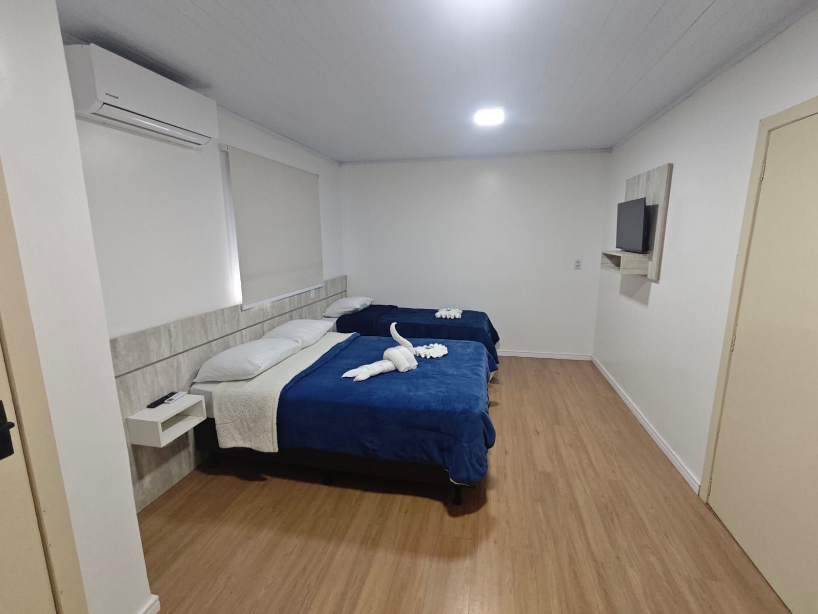 Quarto estilo hotel com duas camas azuis, paredes brancas, piso de madeira e TV fixada na parede.