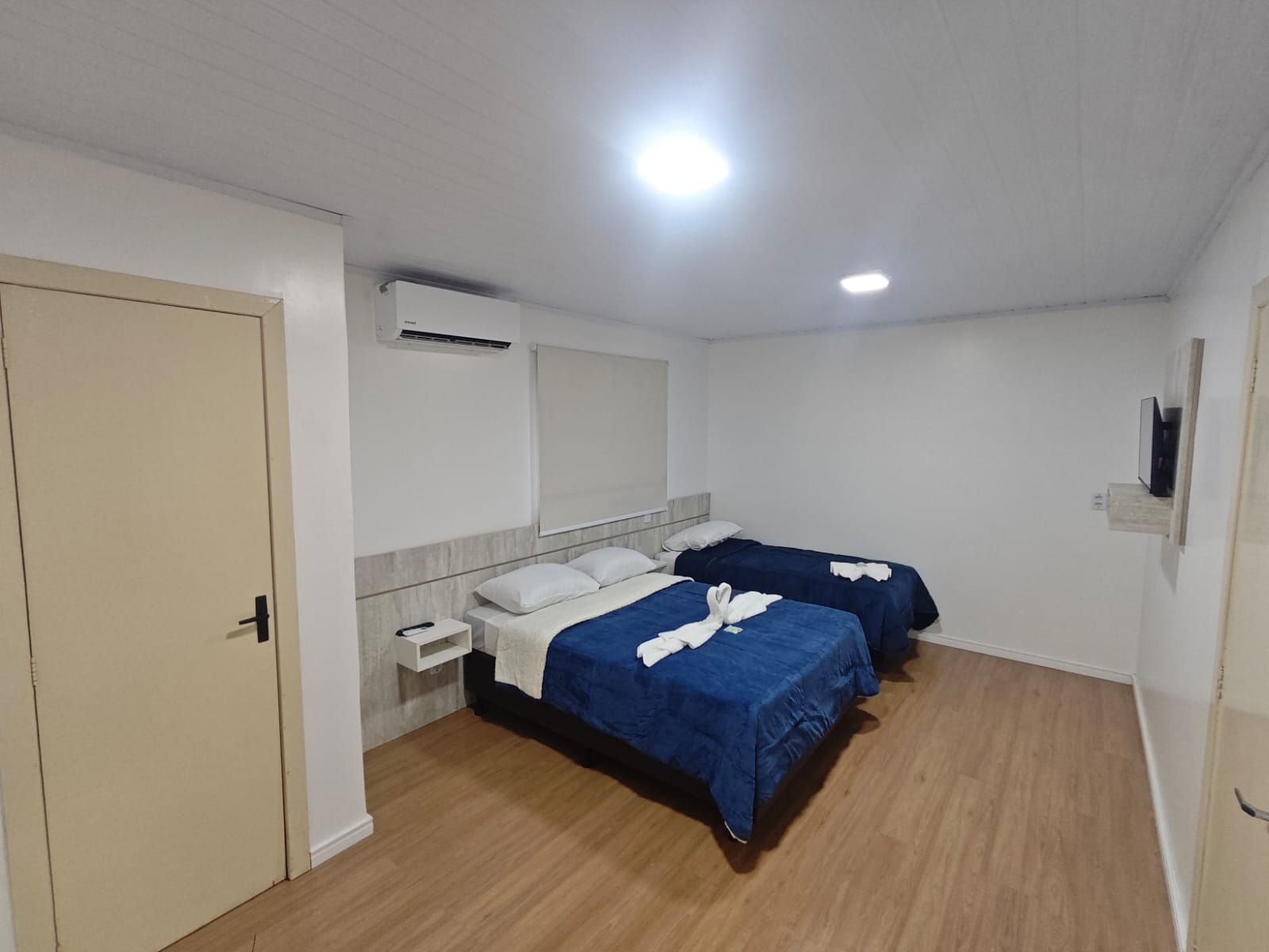 Quarto pequeno com duas camas azuis, paredes brancas, piso de madeira e iluminação embutida no teto.