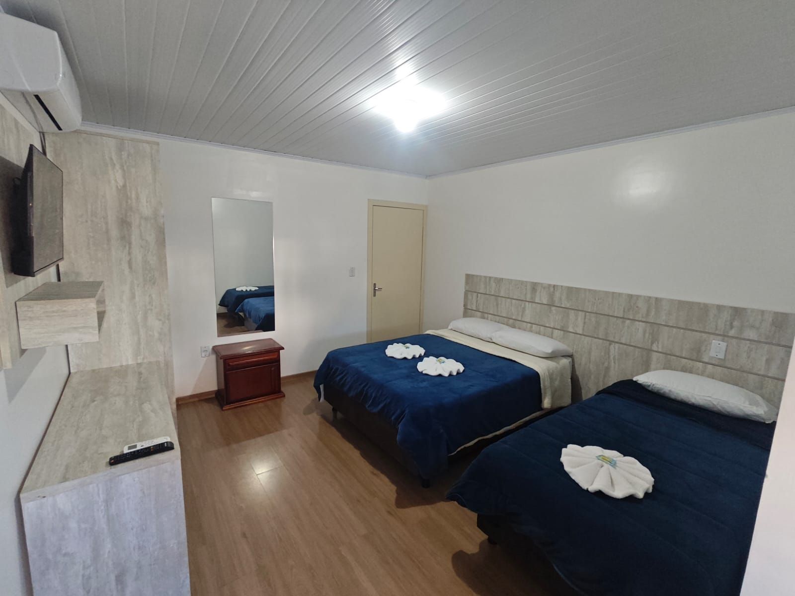Quarto pequeno com duas camas azuis, piso de madeira e paredes brancas sob uma luz brilhante no teto.