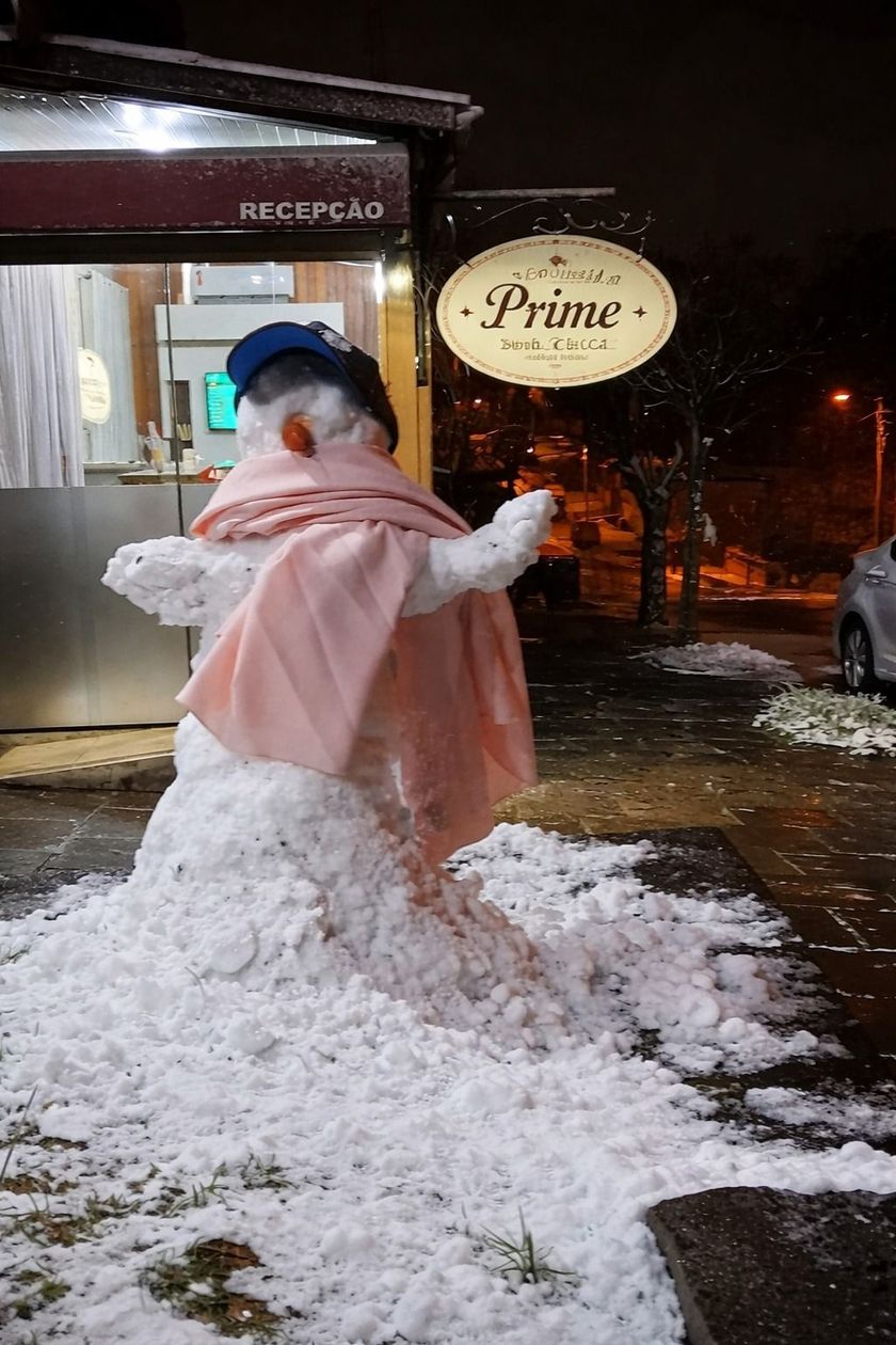 Pessoa com casaco e chapéu rosa em pé na neve em frente a uma loja à noite.