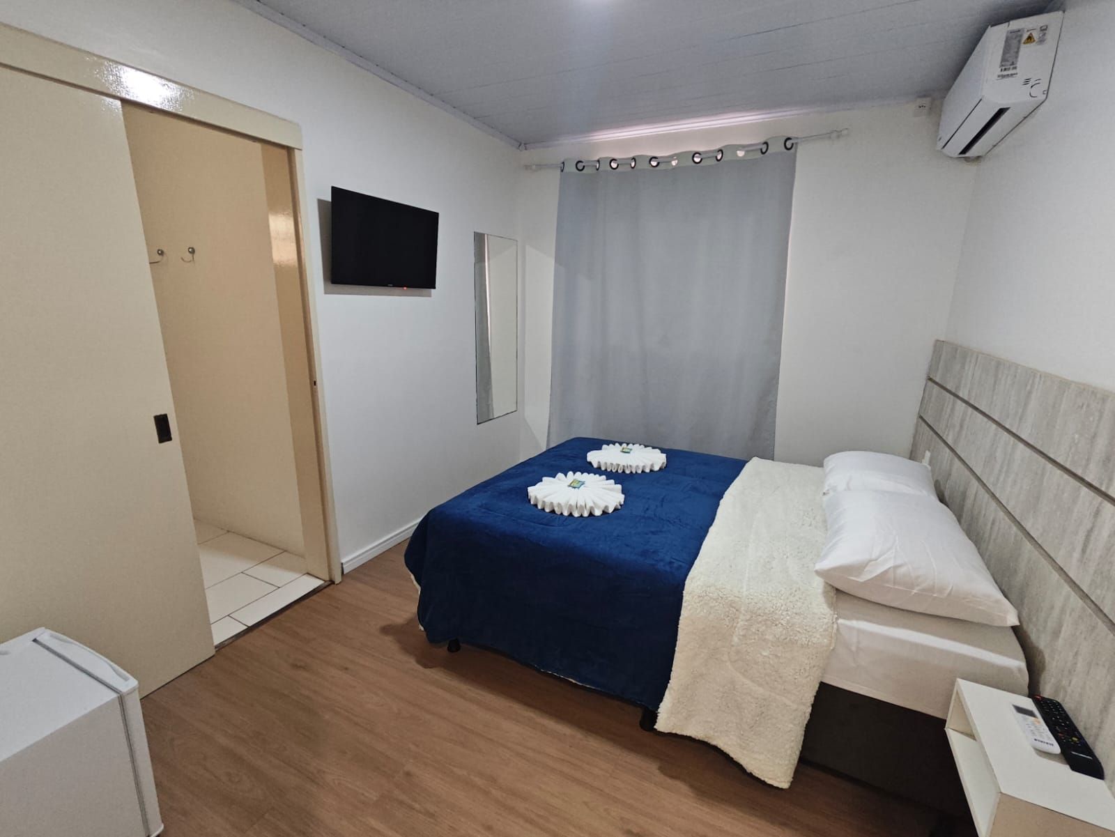 Quarto pequeno com uma cama azul bem arrumada, cortinas brancas, TV de parede e ar condicionado.