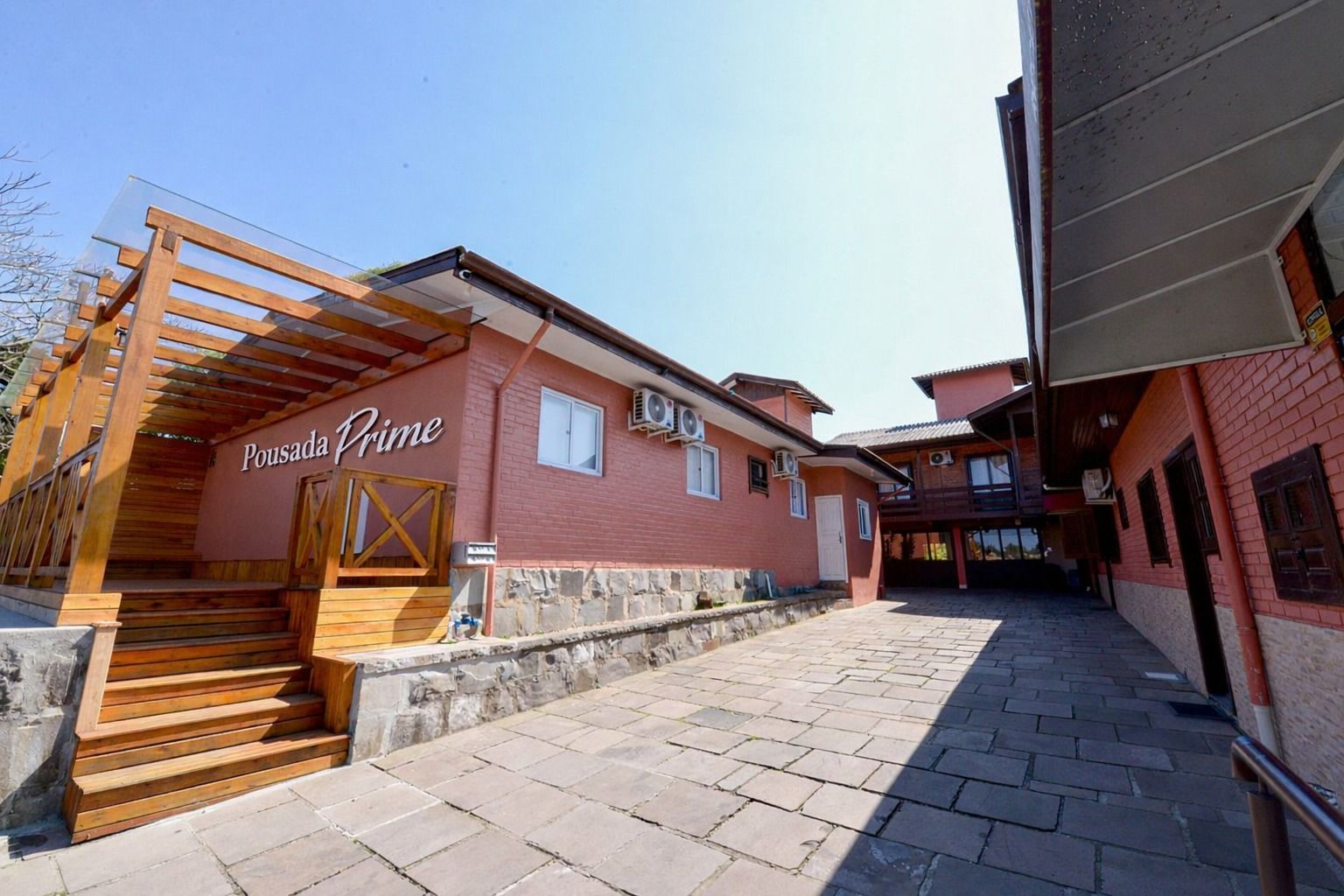 Passarela ensolarada de paralelepípedos entre prédios vermelhos e escadas na entrada de um resort rústico.
