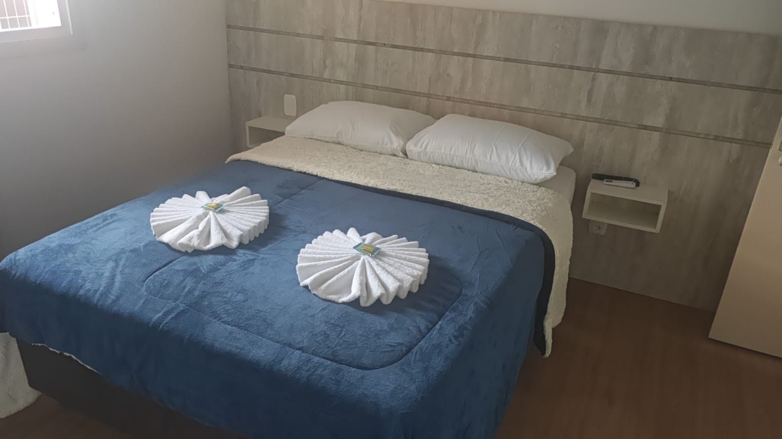 Cama impecavelmente arrumada com cobertor azul, duas toalhas em formato de cisne e travesseiros em um quarto de hotel moderno.