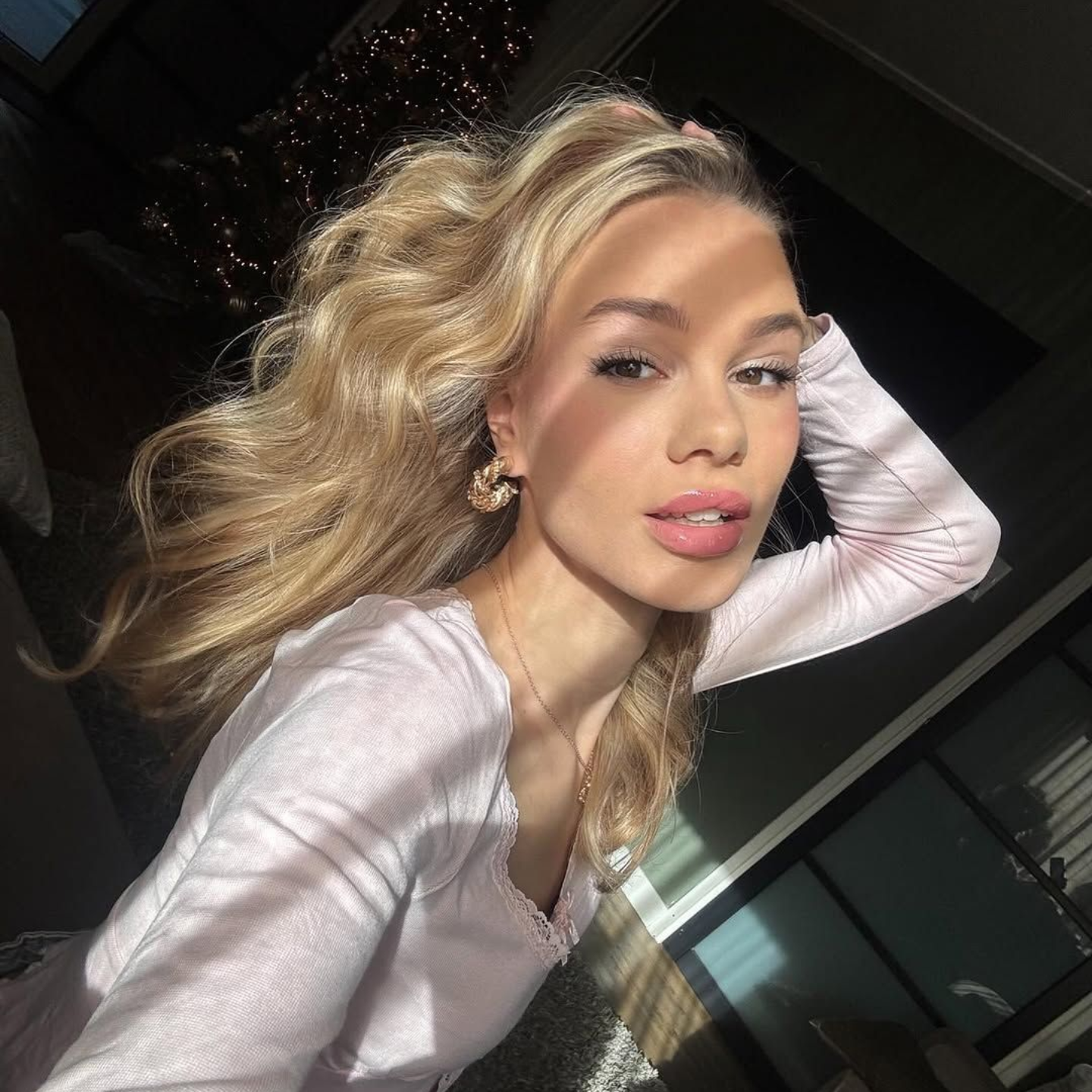 Een persoon met golvend blond haar draagt ββeen lichtgekleurde top en poseert met een hand achter het hoofd in een zonovergoten binnenruimte.