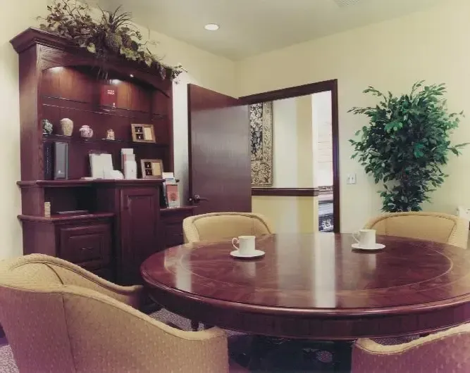 Conference room with round wooden table, beige chairs, and a dark wood cabinet by an open door.