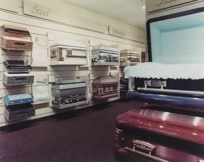 Shelves of assorted caskets in a funeral home showroom with a maroon casket in the foreground