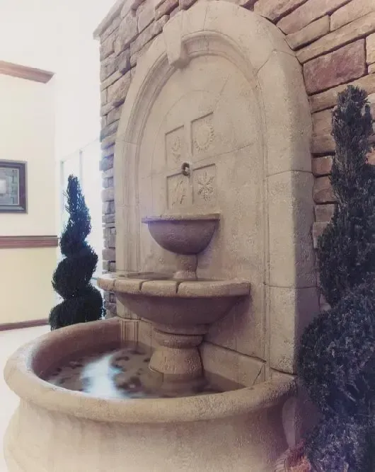 Stone wall fountain with cascading water into a round basin, flanked by tall topiary shrubs.