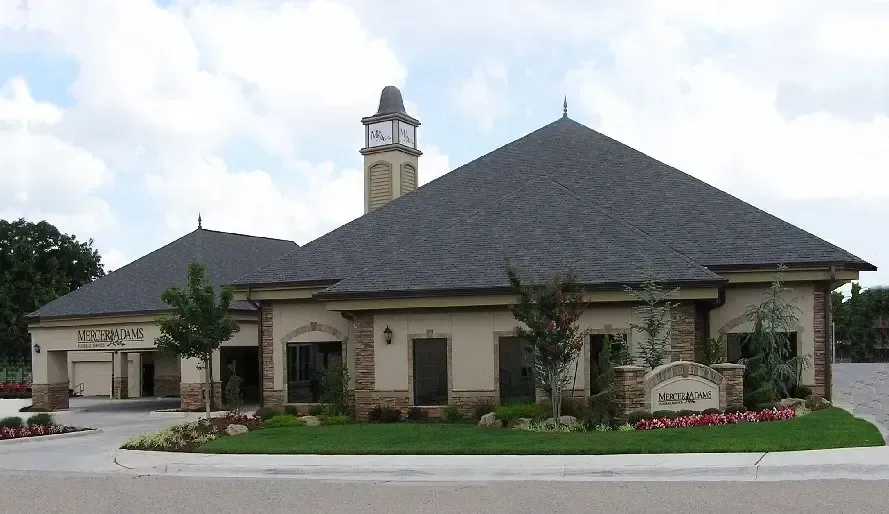 Single-story brick building with a dark roof and stone accents, landscaped entrance, and tower near the corner
