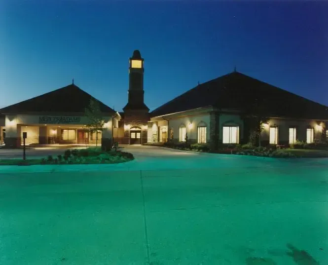 Nighttime view of a lit building with twin peaked roofs and a central tower, reflected in a turquoise courtyard