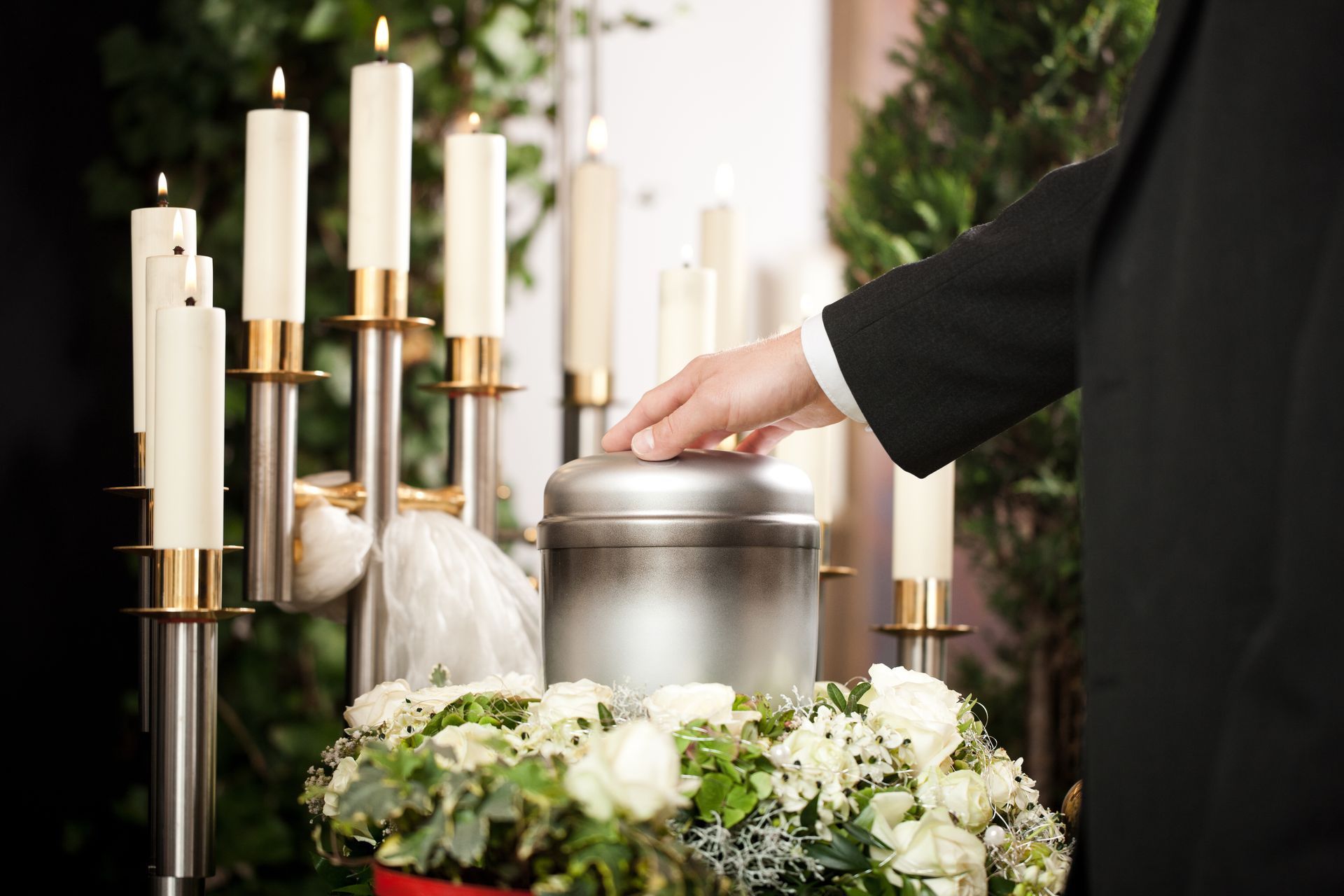 Hand placing a silver urn among white flowers and candles at a memorial altar