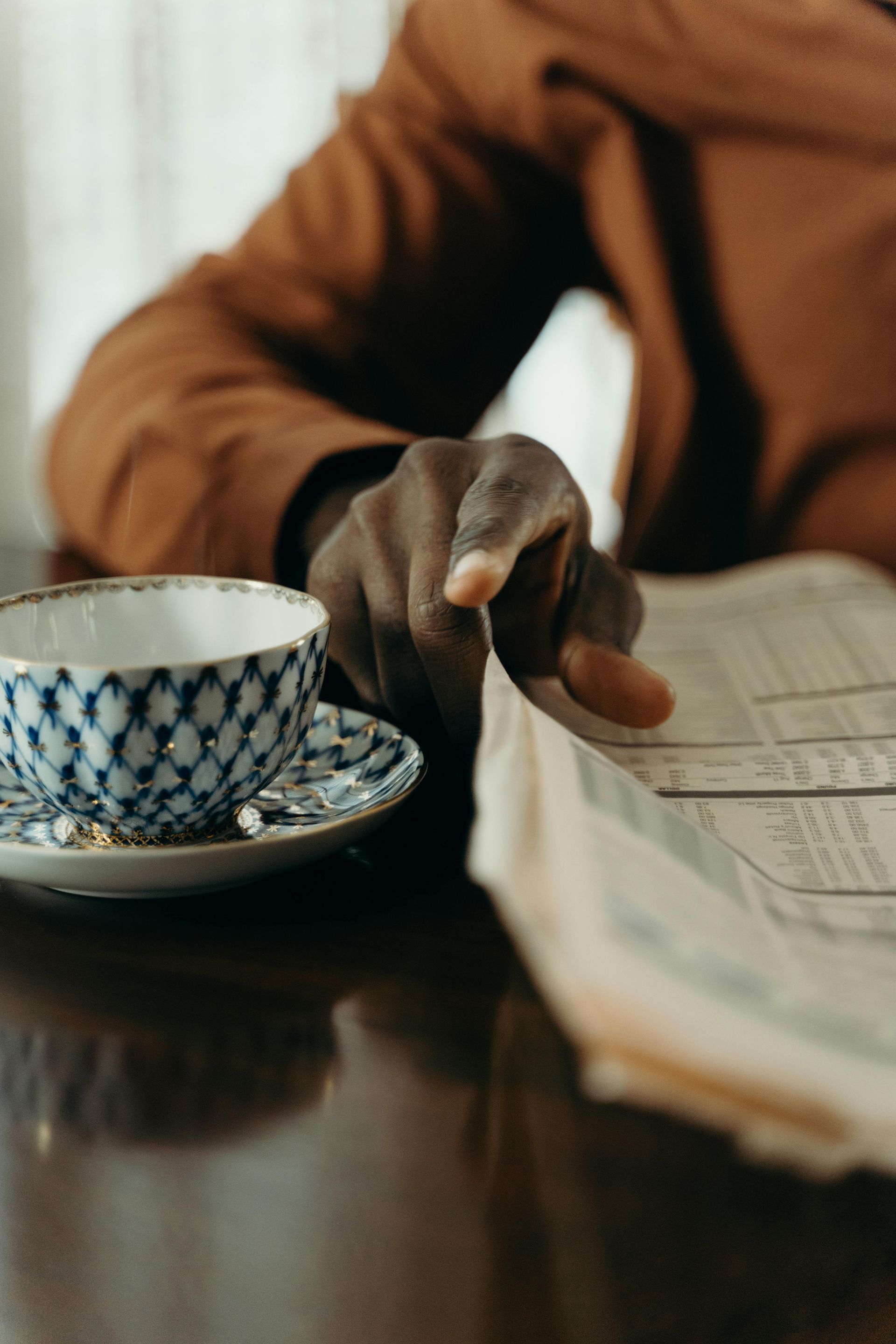 Hand reaching over an open newspaper beside a patterned teacup on a dark table