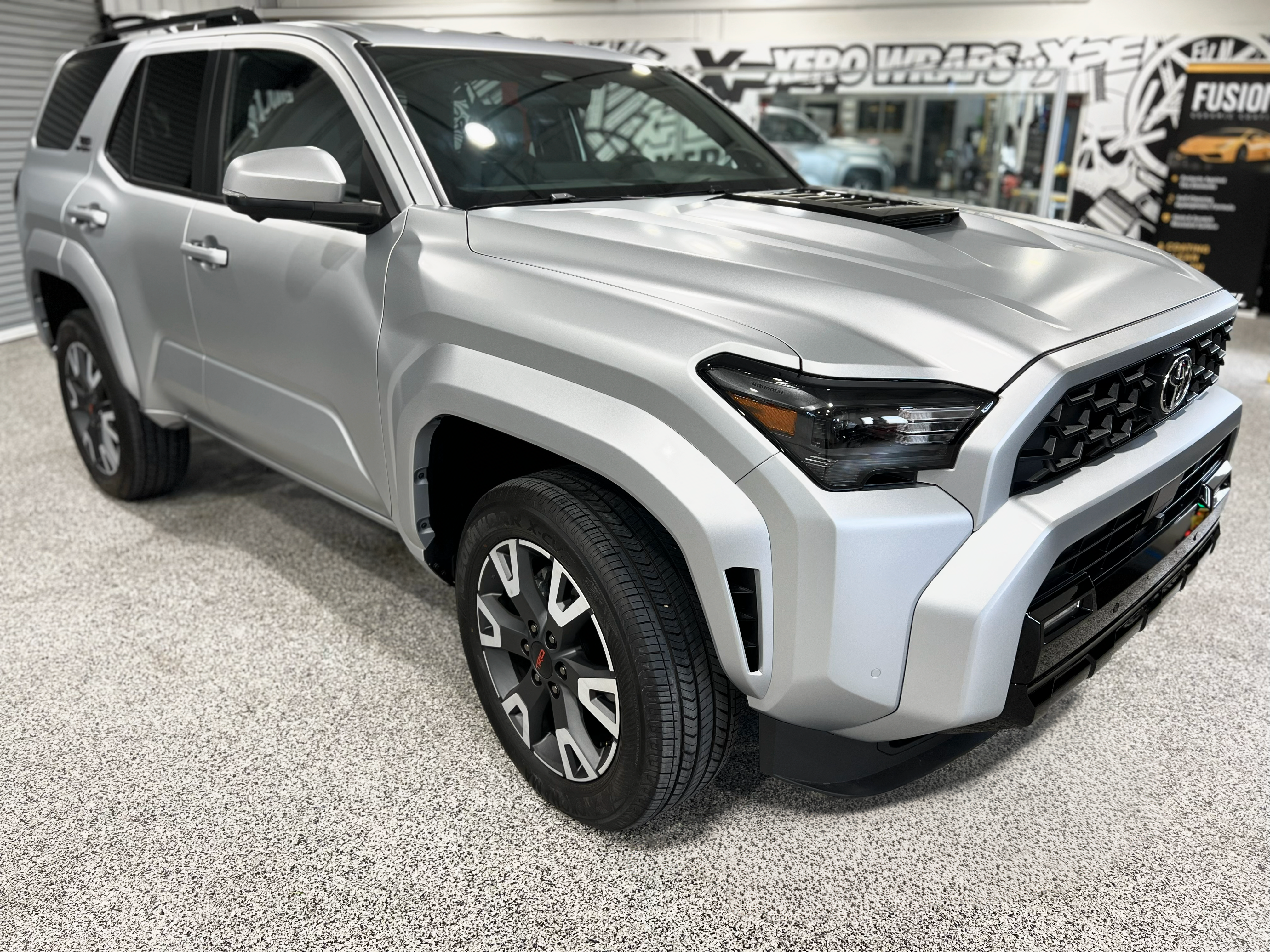 A gray ford bronco is parked in a garage next to a red truck.