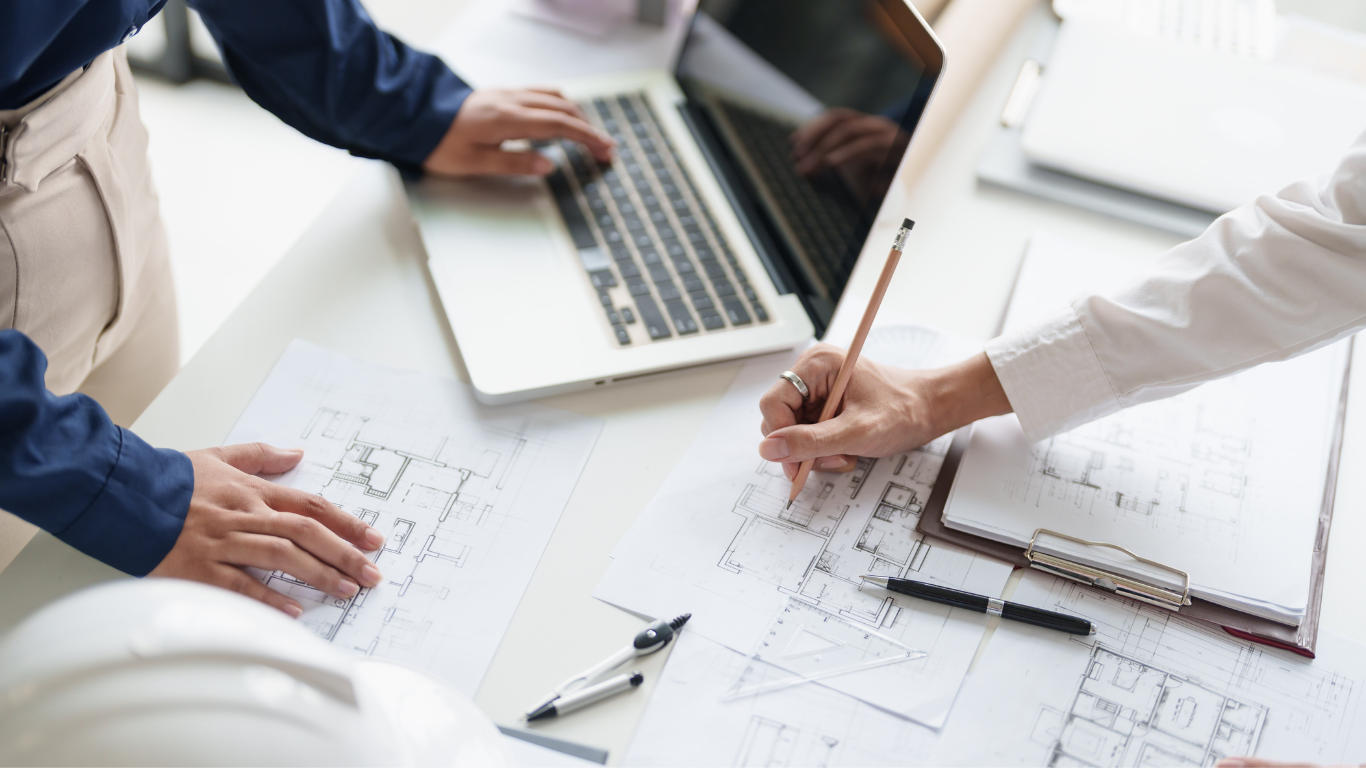 A group of people are sitting at a table with a laptop and blueprints.