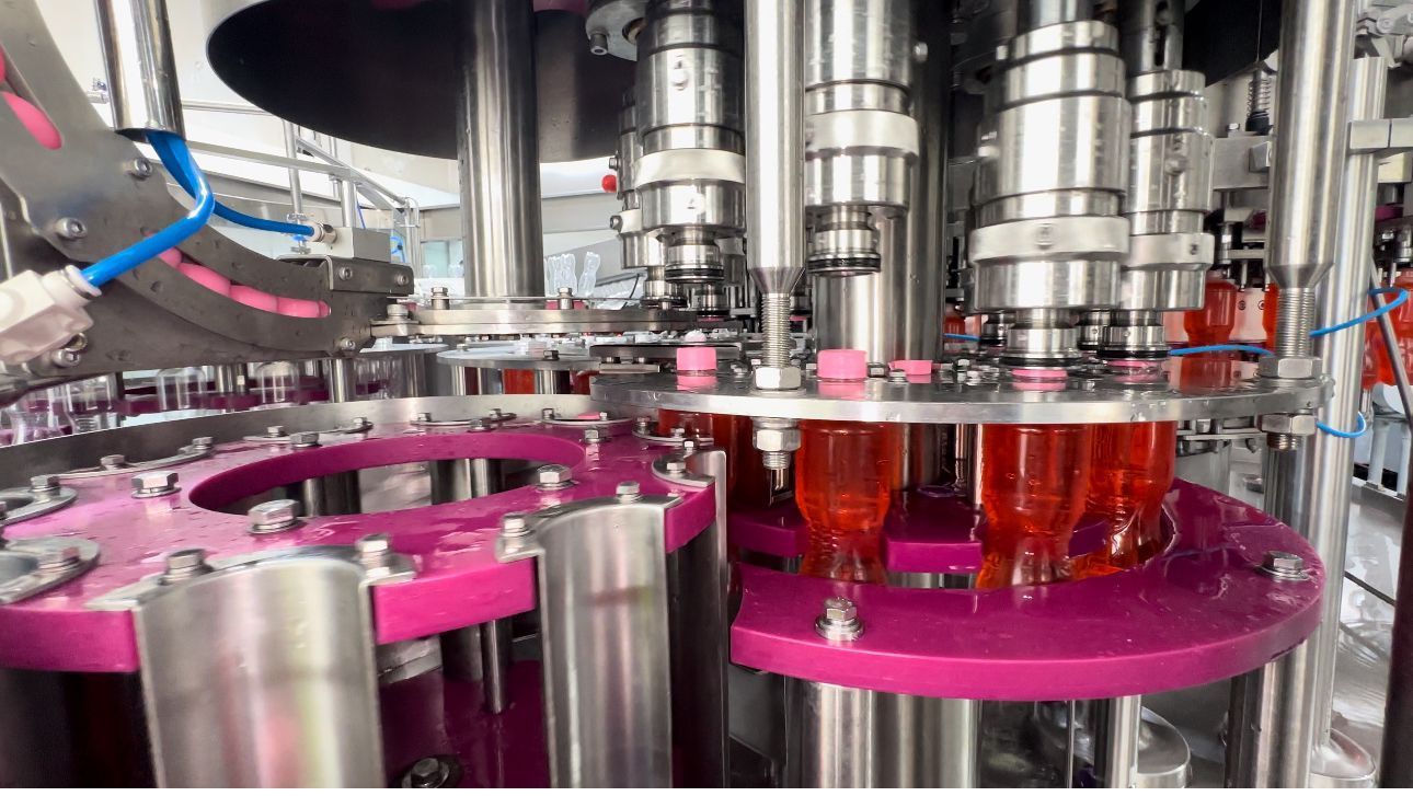 Máquina embotelladora automatizada que llena líquido rosa en botellas transparentes, componentes de acero inoxidable.