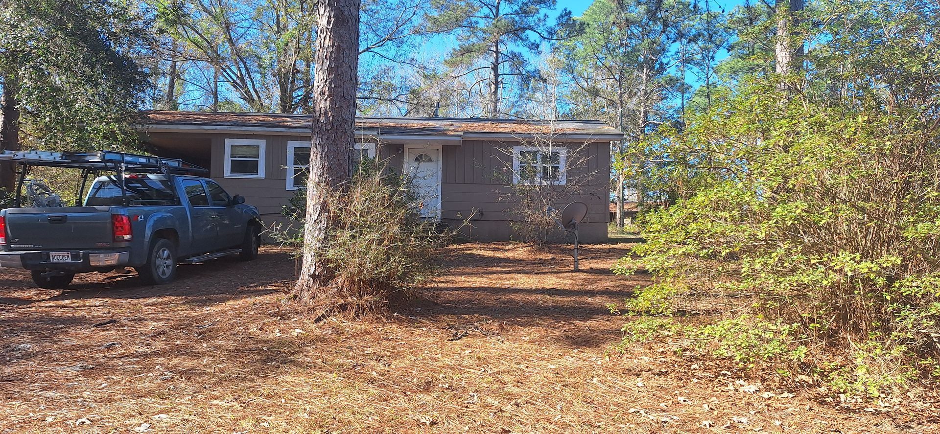 Small brick house with a blue pickup truck parked in the driveway, surrounded by trees and dry leaves.