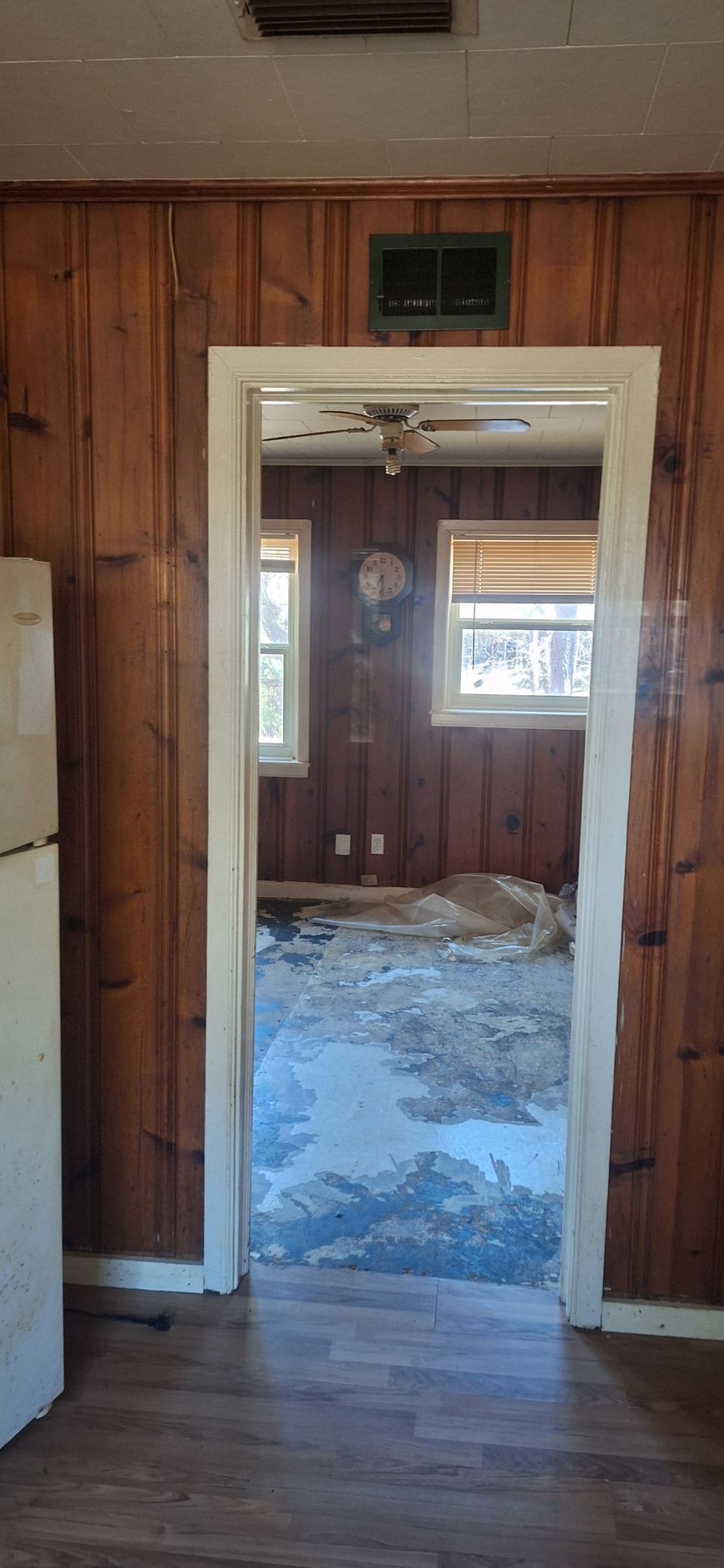 View through a doorway into a wood-paneled room with damaged floor and two windows.