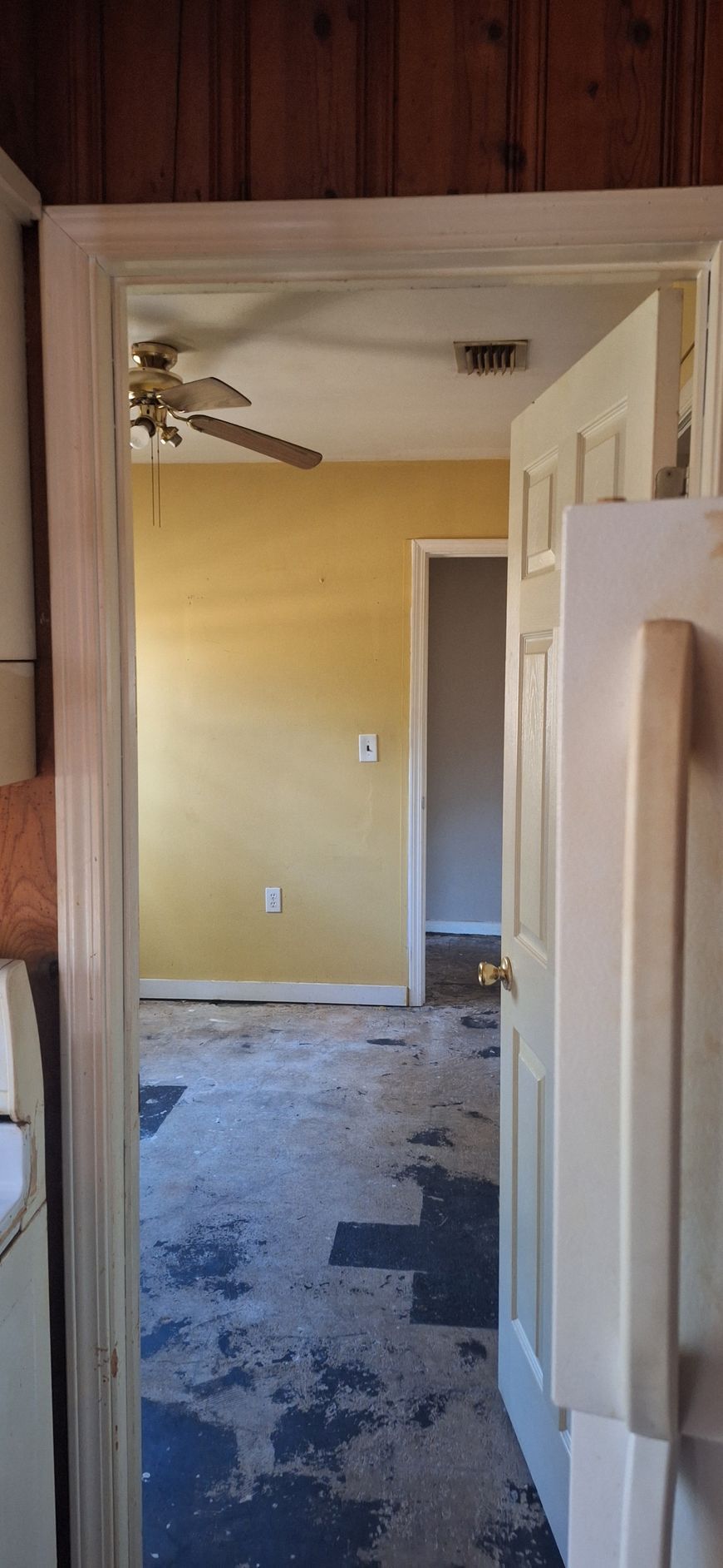 Narrow hallway with wood-paneled wall, open white doors, yellow room ahead, and worn carpet flooring