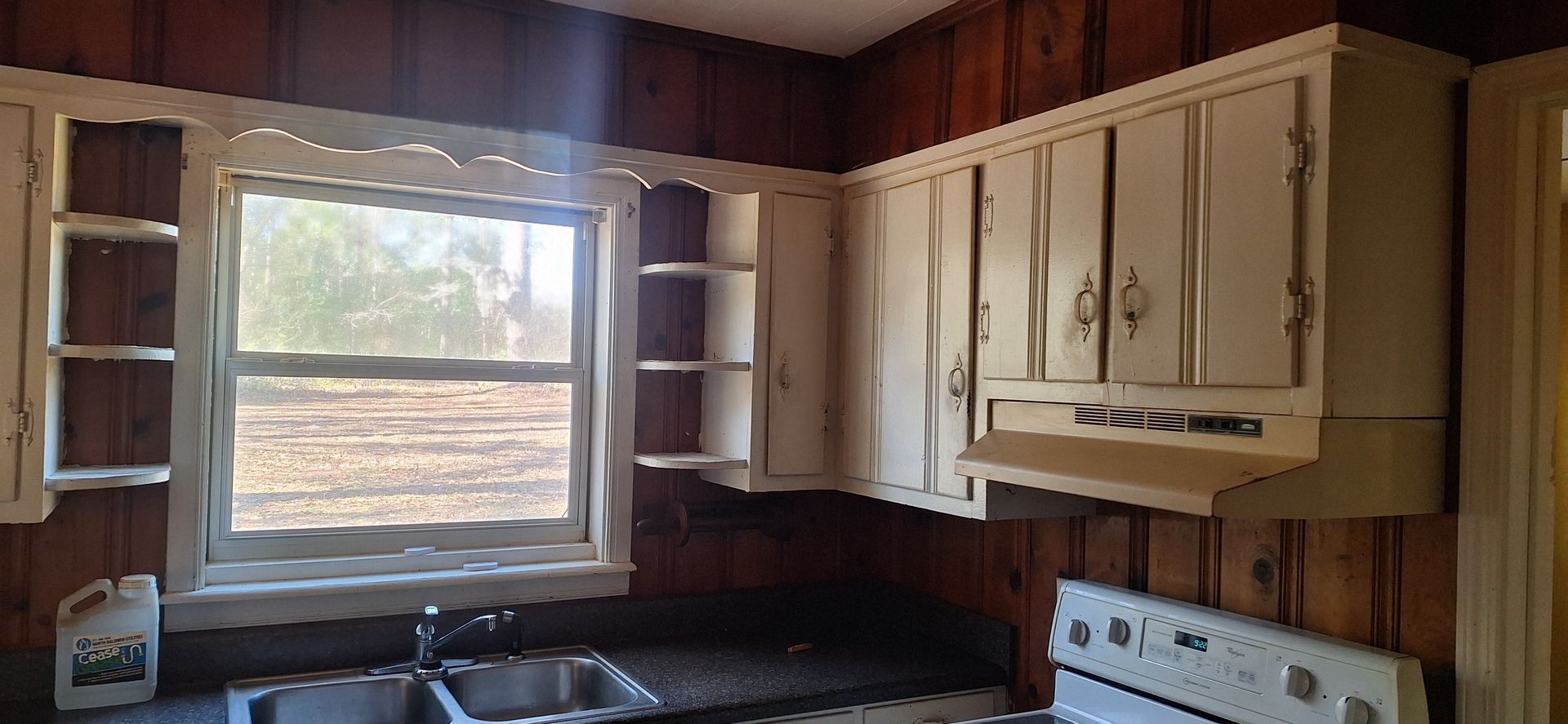 Small rustic kitchen with wood-paneled walls, white cabinets, window, sink, and stove.