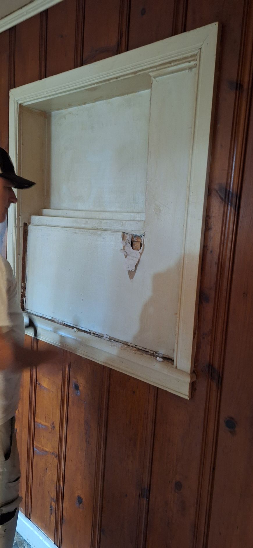Closed white shutter window on wood-paneled wall with a damaged lower panel