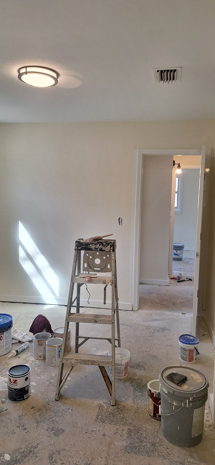 Paint-splattered room under renovation with ladder, paint cans, and doorway to another room