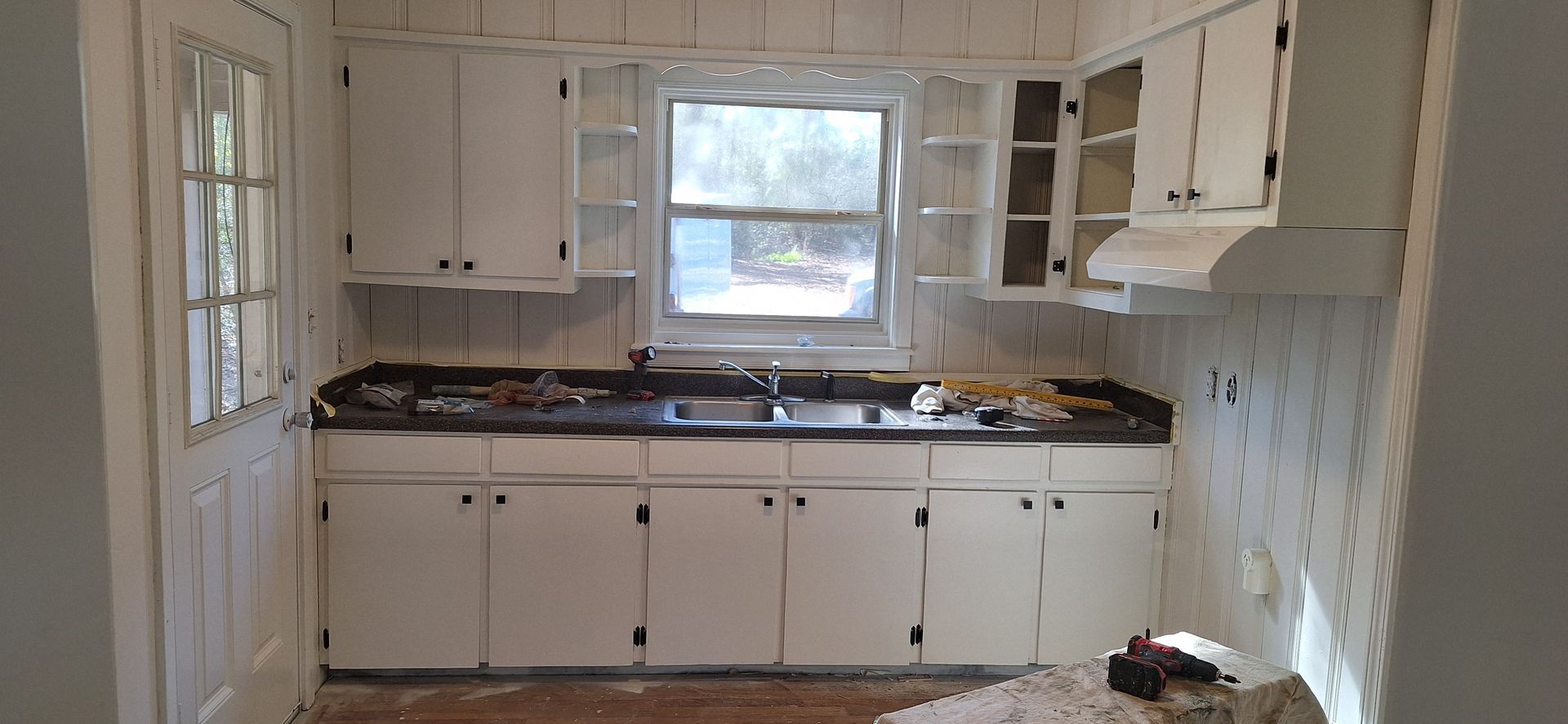 Empty white kitchen under renovation, with cabinets, tiled walls, and a window above the sink.