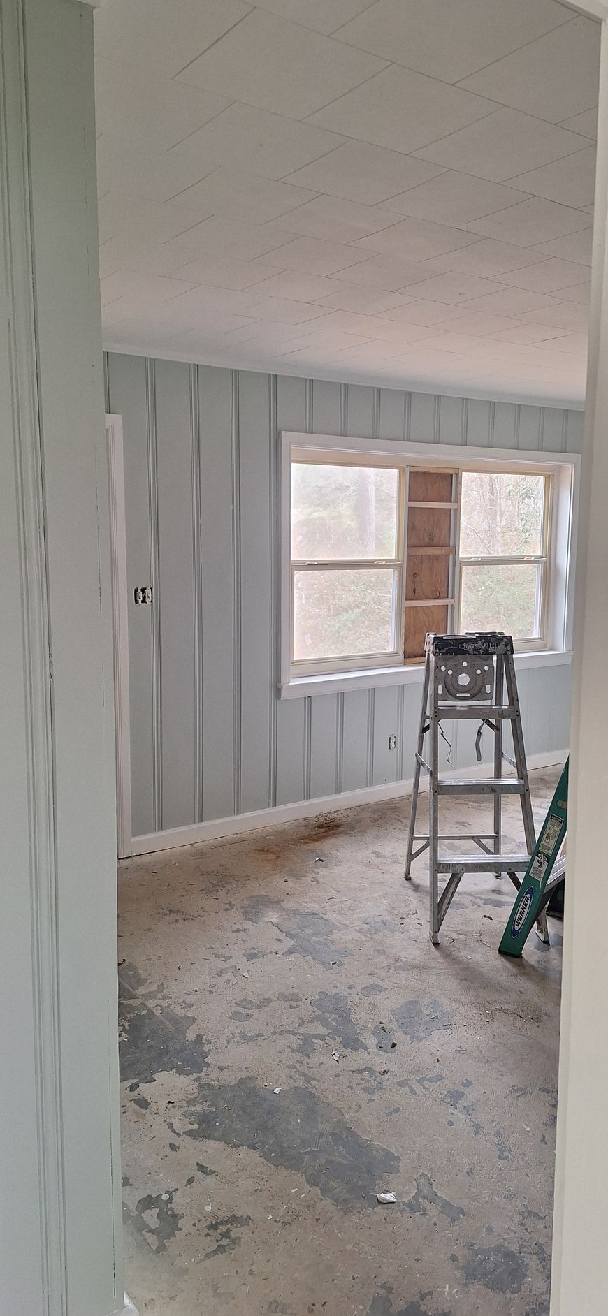 Partially renovated room with gray paneling, a window, ladder, and debris on the floor