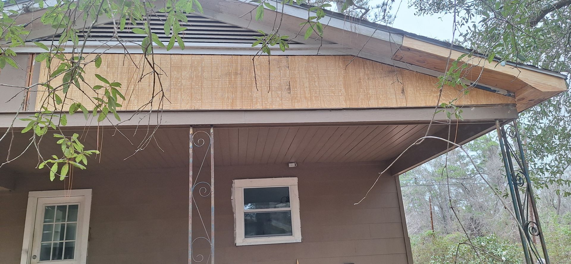 Small brown house facade with a porch roof and windows, framed by tree branches outside.
