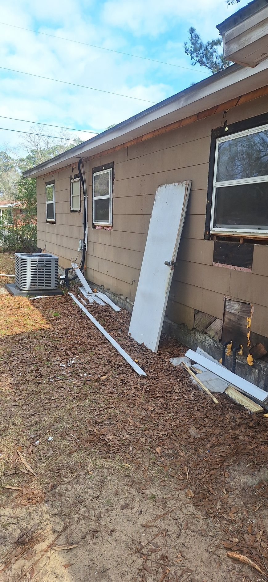 Side of a beige house with a window unit AC, white board leaning against wall, and leaf-covered ground.