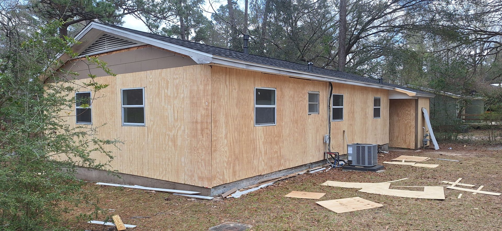 Unfinished tan house with white trim and a metal roof in a wooded yard, with building materials nearby